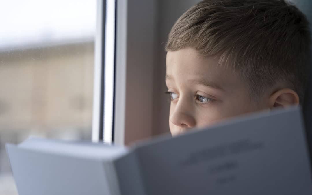 A young boy sitting with a book looking out of the window to signify the signs of dyslexia in a 5 year old